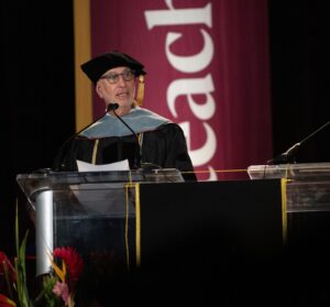 A man in academic regalia speaks at a podium during a graduation ceremony, with a maroon banner in the background.
