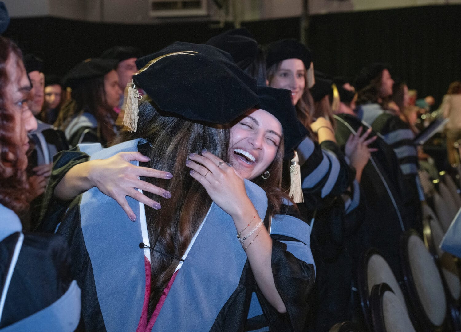 Graduates in caps and gowns embrace and celebrate during a commencement ceremony in an indoor venue.
