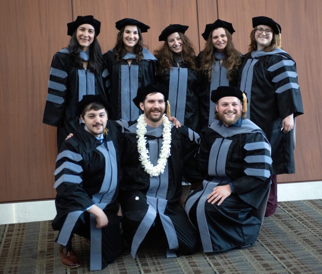 Eight graduates in black academic robes and caps pose for a group photo indoors; one person in front wears a white flower lei.