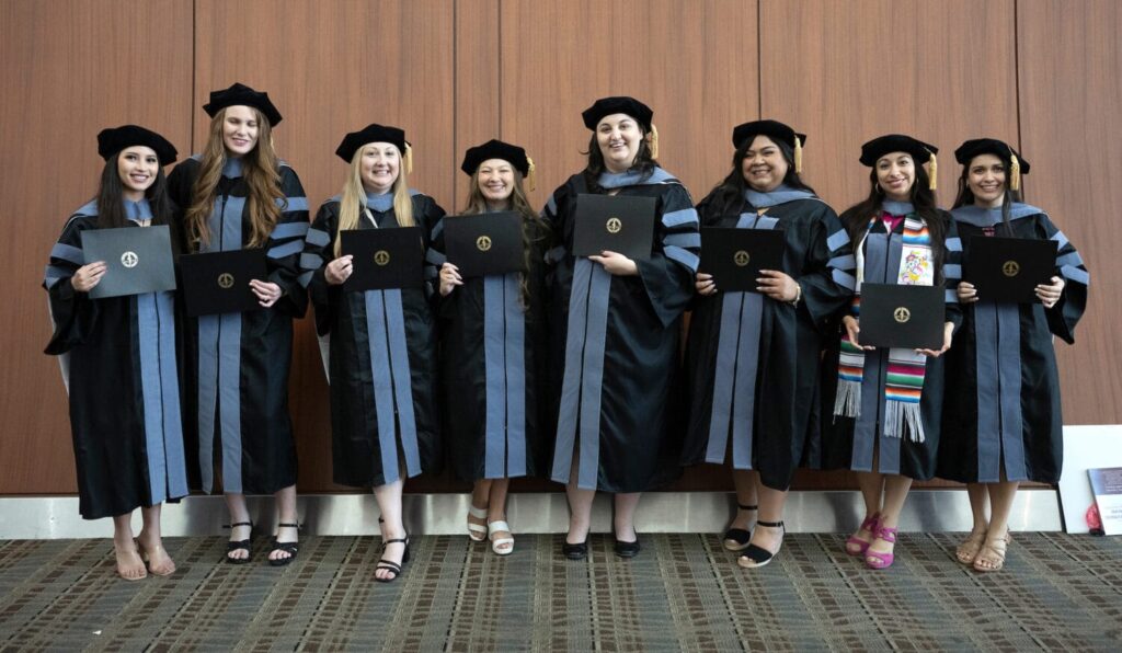 Seven graduates in black academic gowns and caps stand in a row, holding diplomas and smiling in front of a wood-paneled wall.