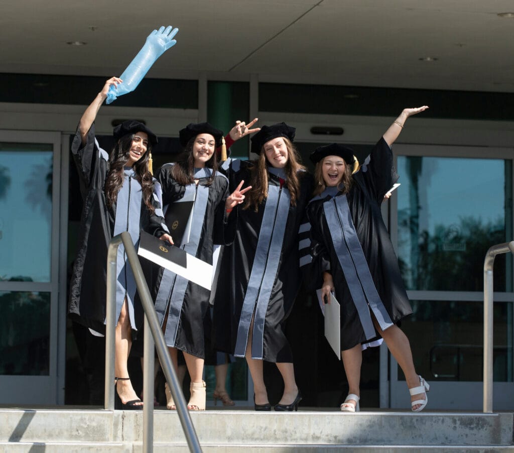 Four graduates in caps and gowns stand on steps, smiling and celebrating. One holds a blue inflatable object. They are outside a building with large glass doors.