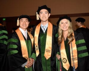 Three people in academic regalia and gold sashes labeled "Podiatric Medicine" smile at a graduation ceremony.