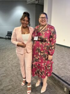 Two women standing indoors, one in a light pink suit and white top, the other in a pink floral dress. They are smiling and posing together on a carpeted floor.