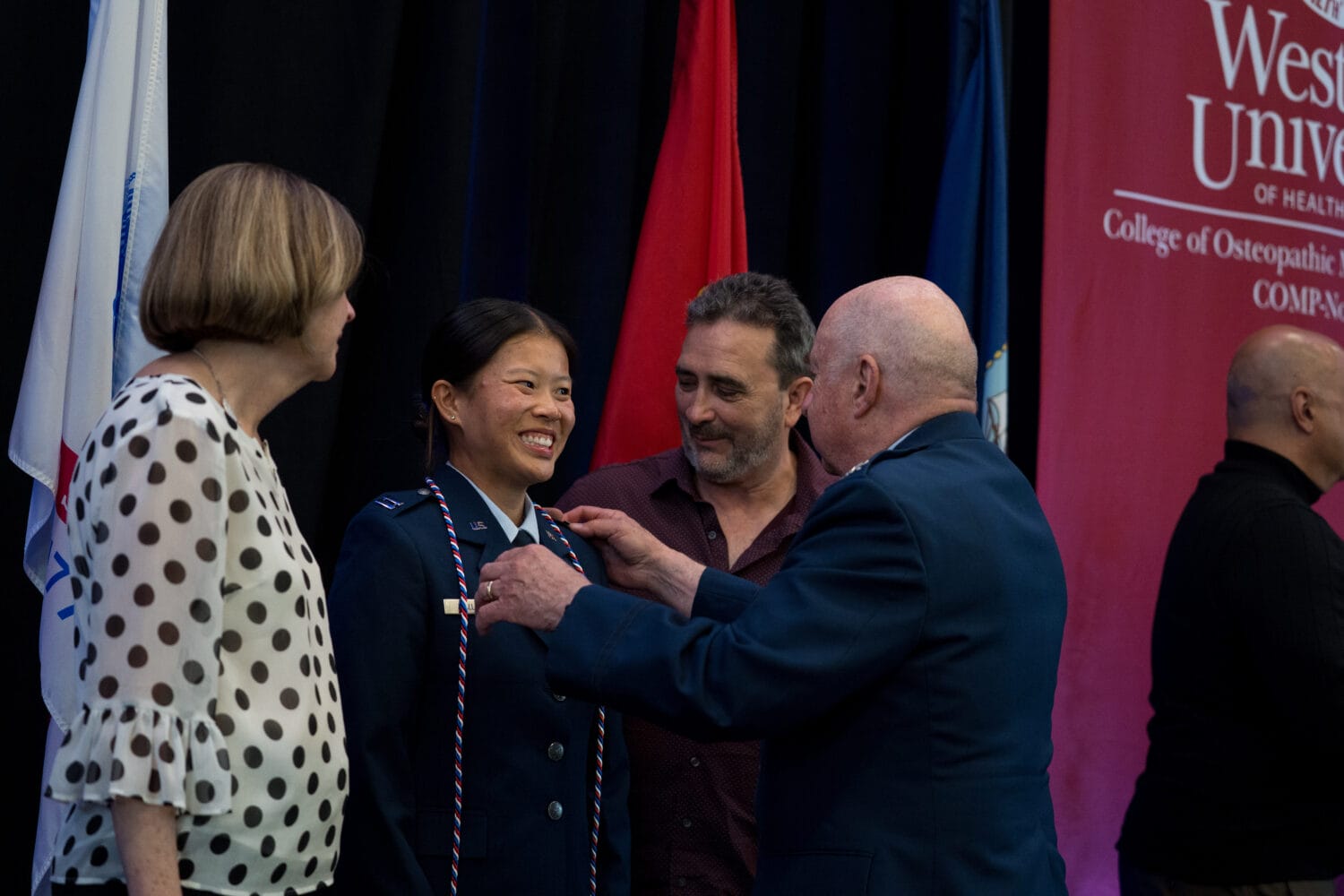 A uniformed woman receives a pin from an older man at a ceremony, with two other people standing nearby and university banners in the background.