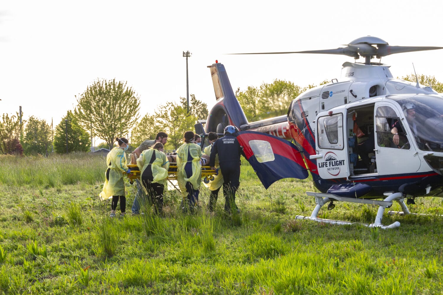 A group of emergency responders in yellow gowns carry a person on a stretcher toward a Life Flight helicopter in a grassy field.