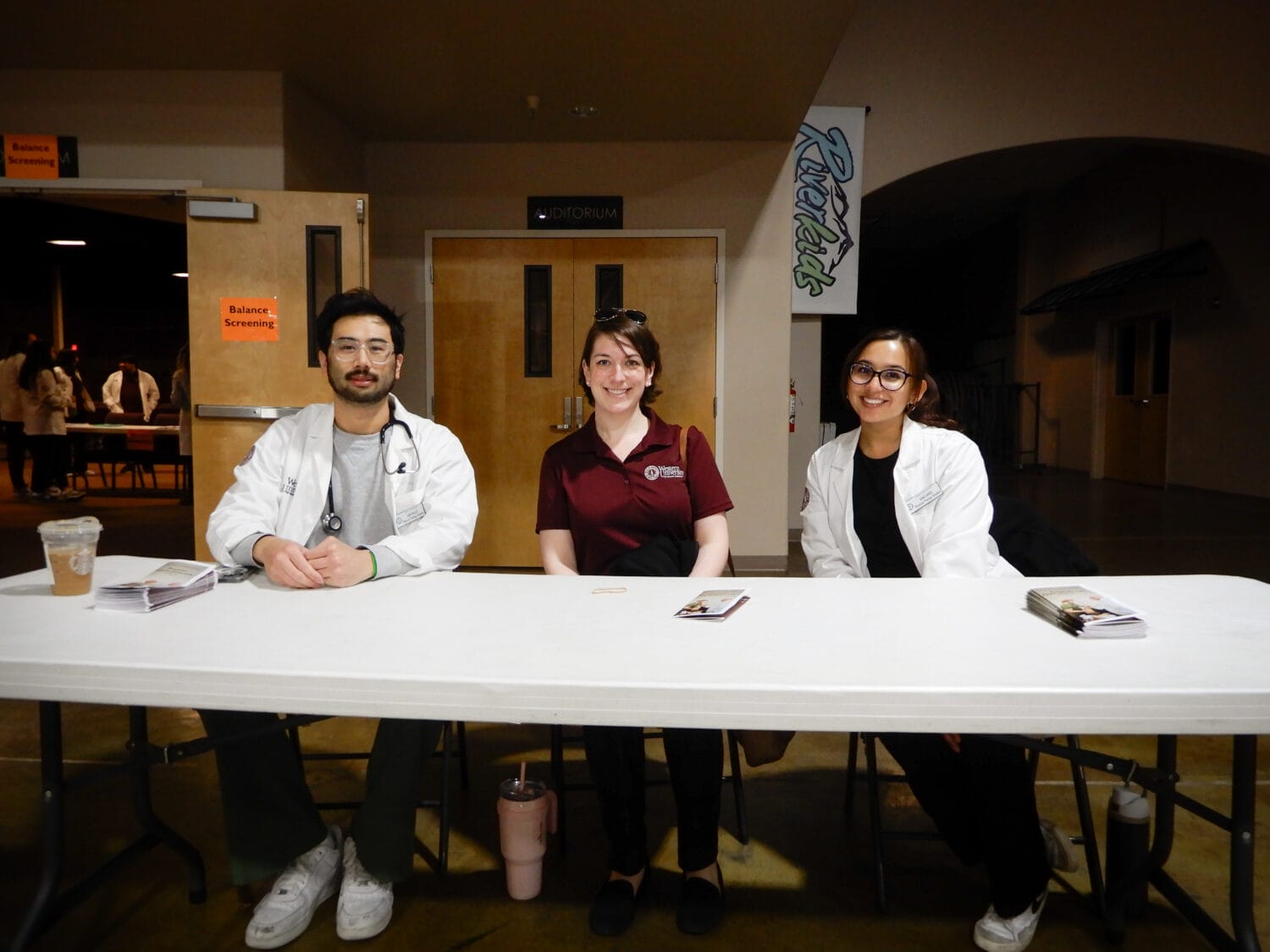 Three people sit behind a white table with papers and drinks in front of them in a well-lit indoor setting, possibly at an event check-in or registration area.