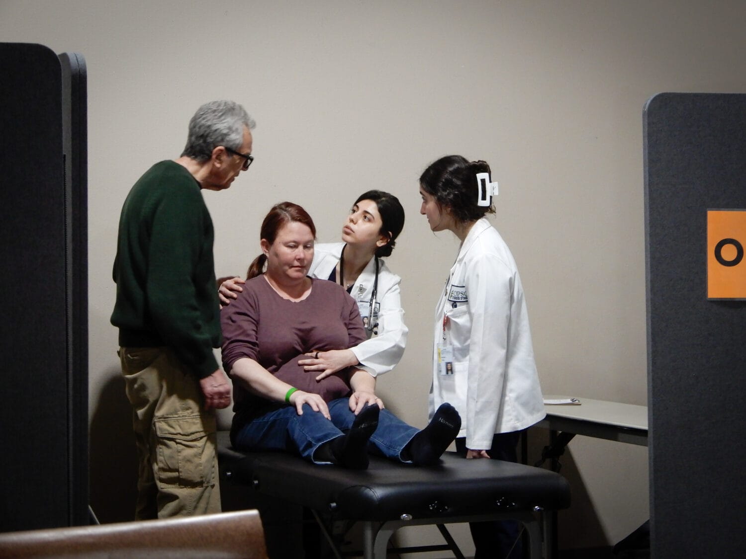 A woman sits on a medical exam table while two healthcare professionals and another adult stand nearby, engaging with her during a consultation.
