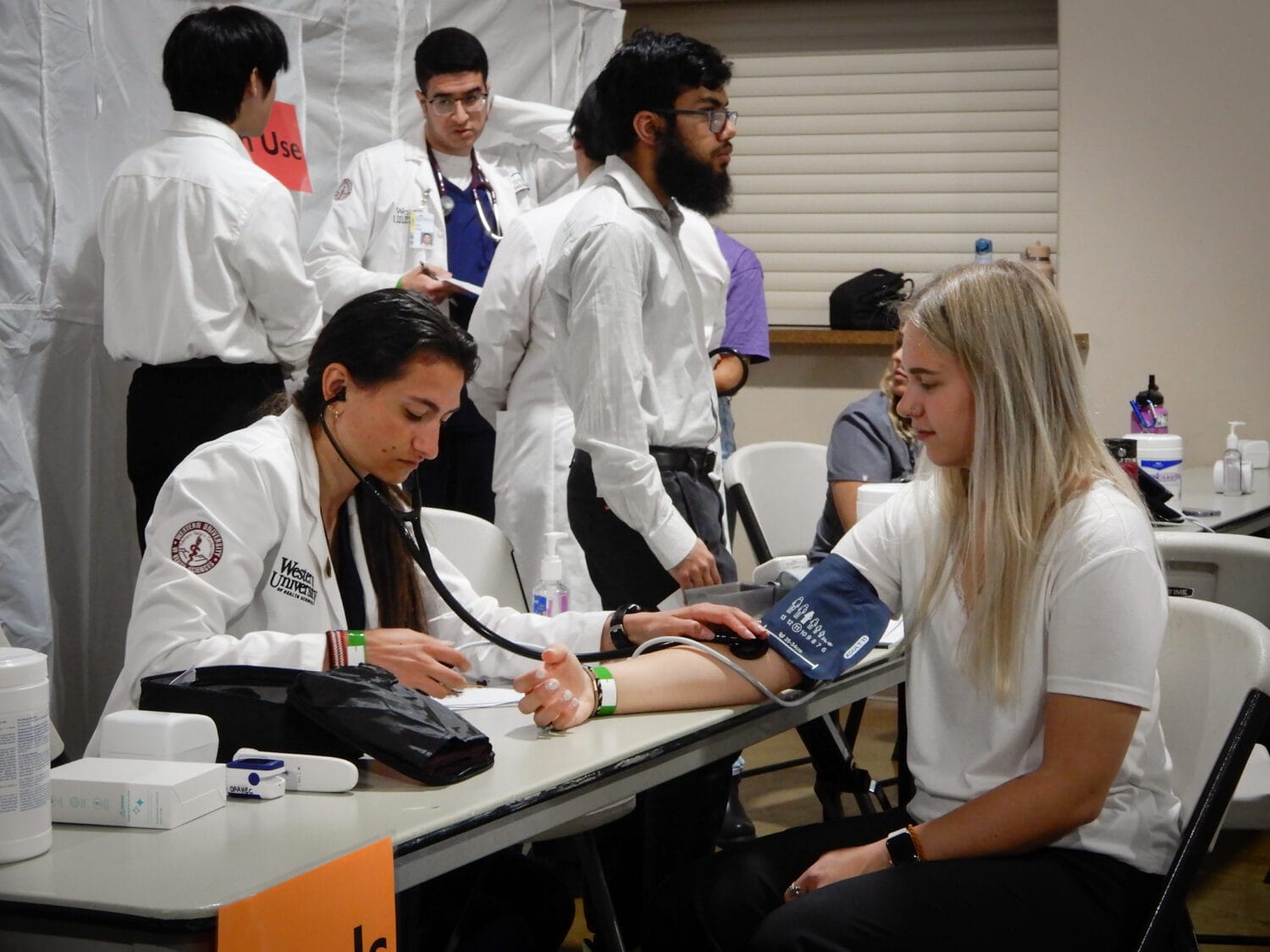 A healthcare professional measures a seated woman's blood pressure at a clinic, while others work in the background.