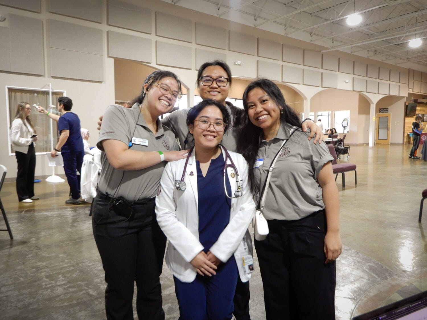 Four smiling people, three in grey uniforms and one in a white coat with a stethoscope, pose together indoors at an event or clinic.