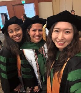 Three graduates in academic regalia smile for a selfie at a graduation ceremony indoors.