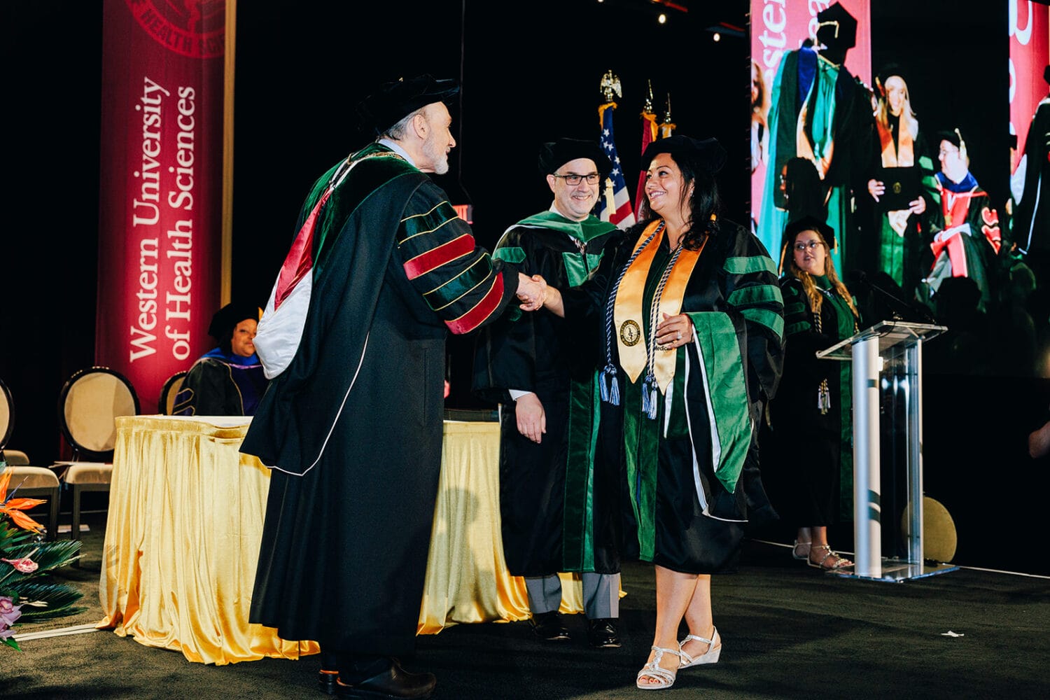 A graduate in cap and gown shakes hands with a faculty member on stage during a Western University of Health Sciences graduation ceremony. Other faculty and staff look on.