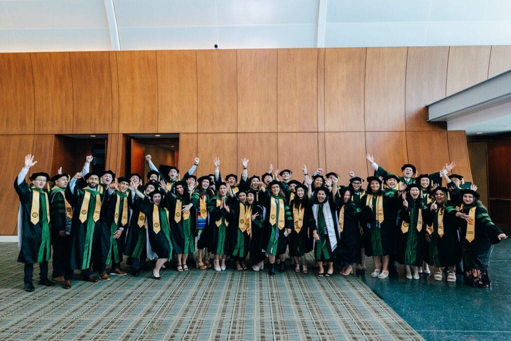 A large group of graduates in green and gold academic gowns and caps pose indoors, some with hands raised, in front of a wood-paneled wall.