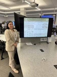 A woman in a beige suit stands next to a monitor displaying a scientific research poster in a conference room labeled "Table 15.