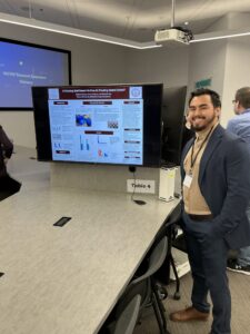 A man stands next to a scientific research poster displayed on a large monitor at a symposium, with other attendees visible in the background.