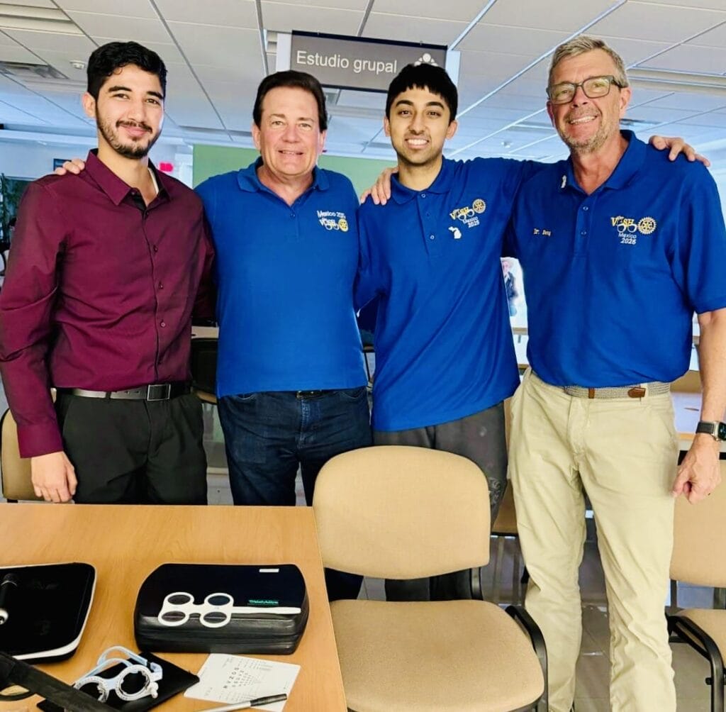 Four men stand together indoors, three wearing blue polo shirts with logos and one in a burgundy shirt, posing and smiling near a table with electronic devices.