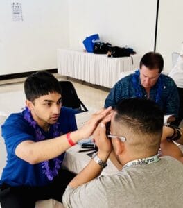A man in a blue shirt examines another man's eyes in a clinical setting while another person writes notes in the background.