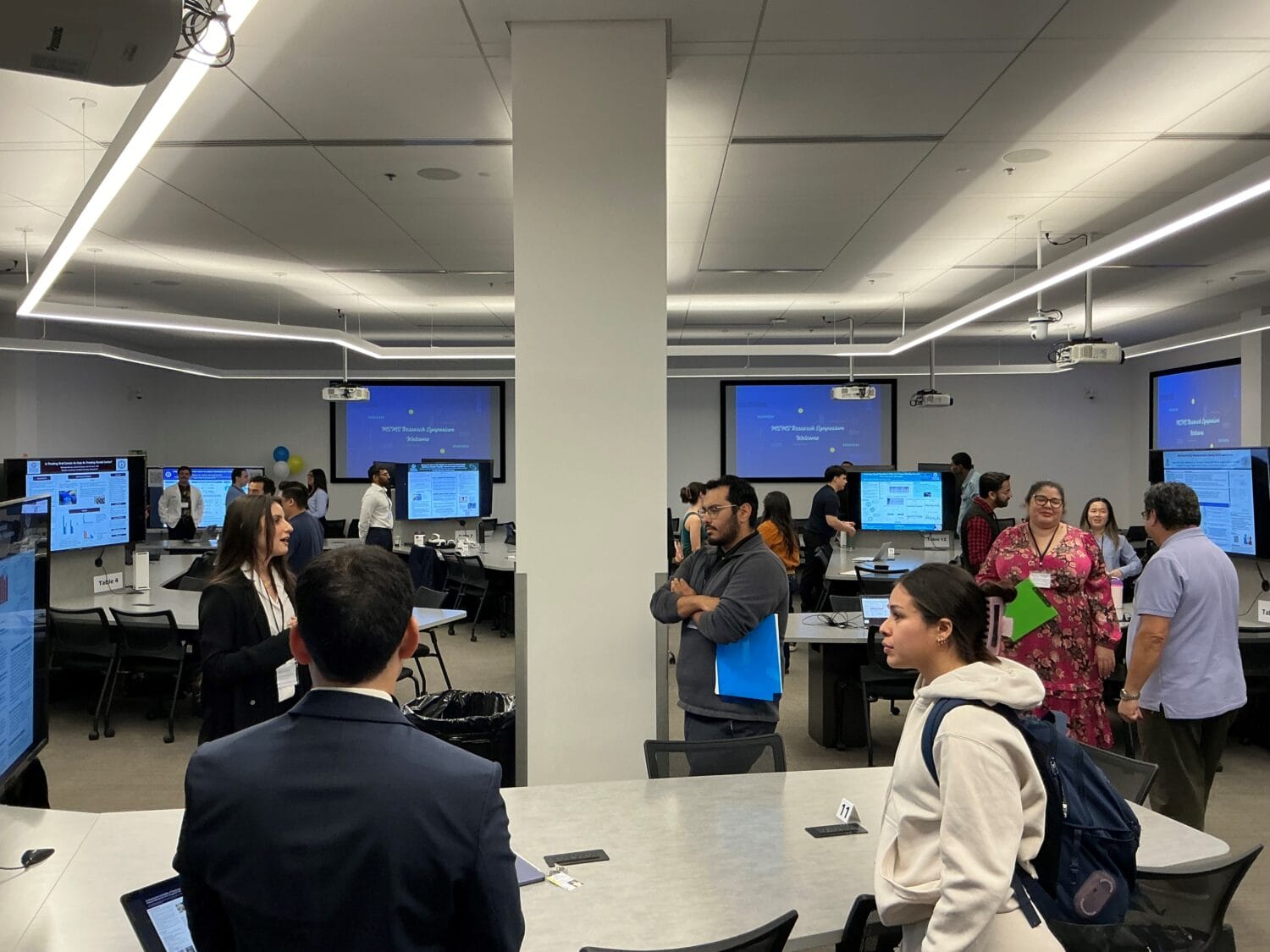 People interact and view displays in a modern conference room with multiple screens showing presentations. Tables and chairs are arranged throughout the space.