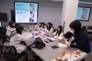 A group of students sits around a table making crafts with beads and paper, while an instructor assists; “Activity Time!” instructions are displayed on screens in the background.
