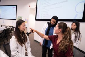 A medical instructor demonstrates an eye exam on a student while two other students observe in a classroom with medical slides on the screens.