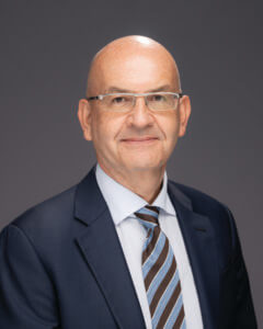 A man wearing glasses, a dark blue suit, white shirt, and striped tie, posing for a professional studio portrait against a plain dark background.