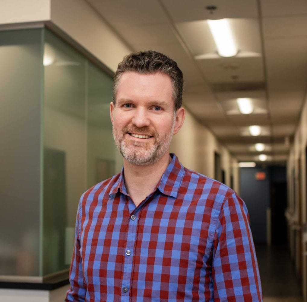 A man with short curly hair and a beard, wearing a blue and red plaid shirt, stands and smiles in a hallway with offices and fluorescent lighting.
