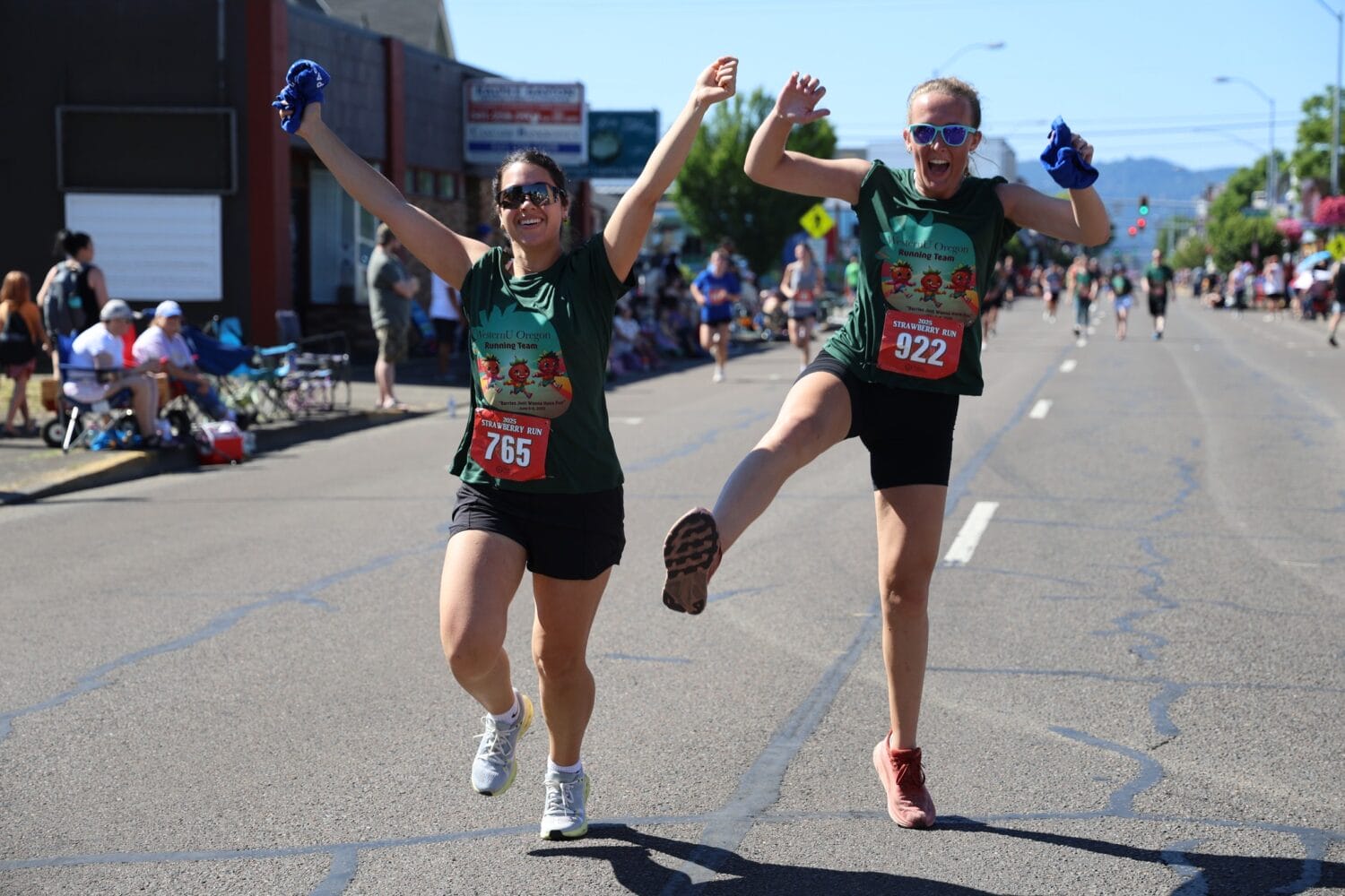 Two women in green shirts and race bibs run and celebrate on a sunny street during a race event, surrounded by other participants and spectators.