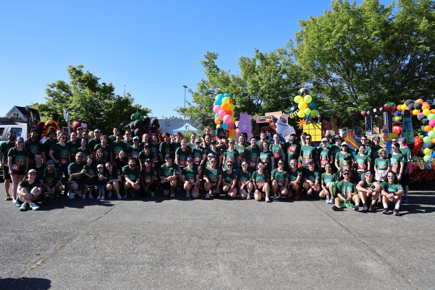 A large group of people in matching green shirts pose for a photo outdoors in front of parade floats decorated with colorful balloons and trees in the background.