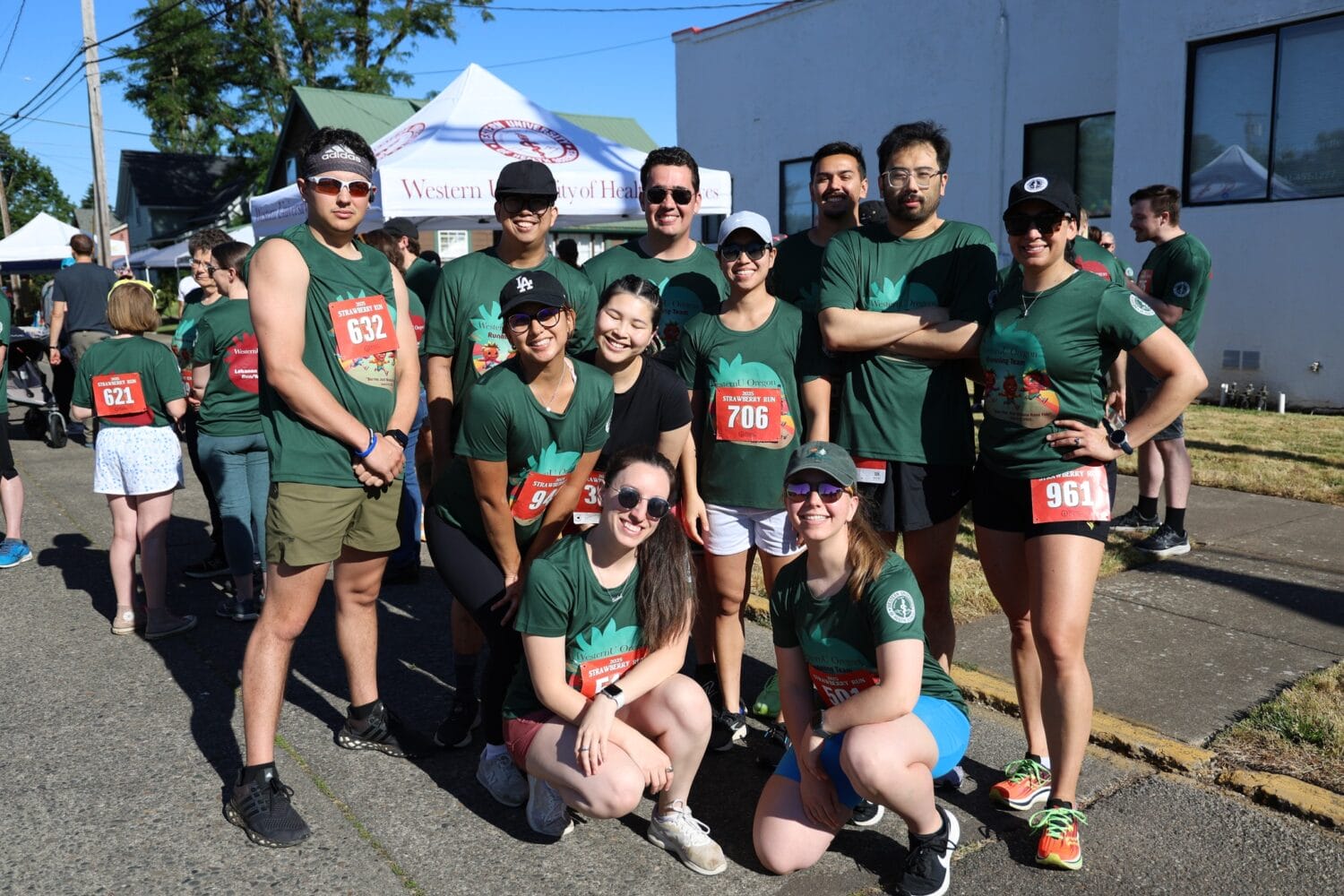 A group of runners wearing green shirts and race bibs pose together outdoors at a running event on a sunny day.