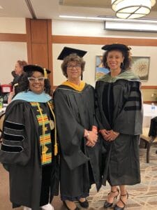 Three women in academic regalia stand together and smile inside a room with brown and white walls, artwork, and a table in the background.