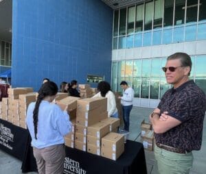 People stand around tables stacked with cardboard boxes outside a building labeled "Western University." One person stands in the foreground wearing sunglasses.