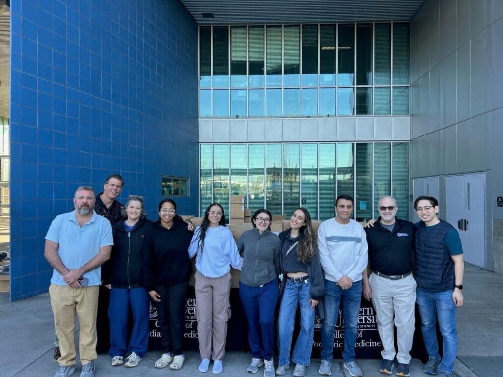 A group of eleven people stands in a row outside a modern building with blue-tiled walls and large windows, smiling at the camera.