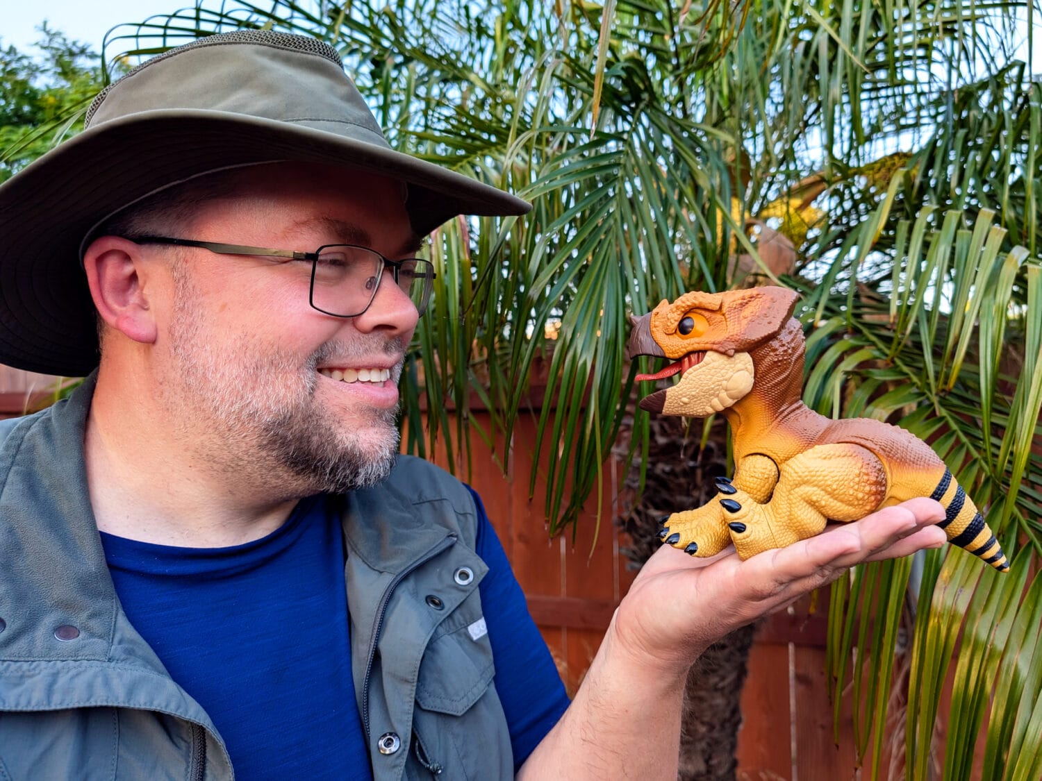 A man wearing glasses and a wide-brim hat smiles while holding a small toy dinosaur in his hand, with palm trees and a wooden fence in the background.