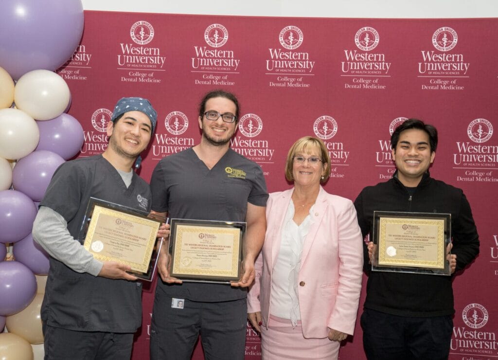 Three people in scrubs hold certificates and stand next to a woman in a pink suit, posing in front of a Western University College of Dental Medicine backdrop and balloon decorations.