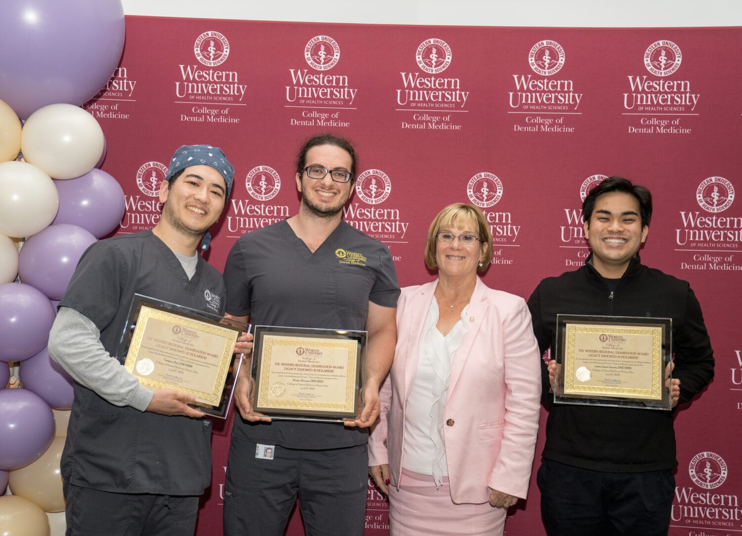 Three people in scrubs hold certificates and stand next to a woman in a pink suit, posing in front of a Western University College of Dental Medicine backdrop and balloon decorations.