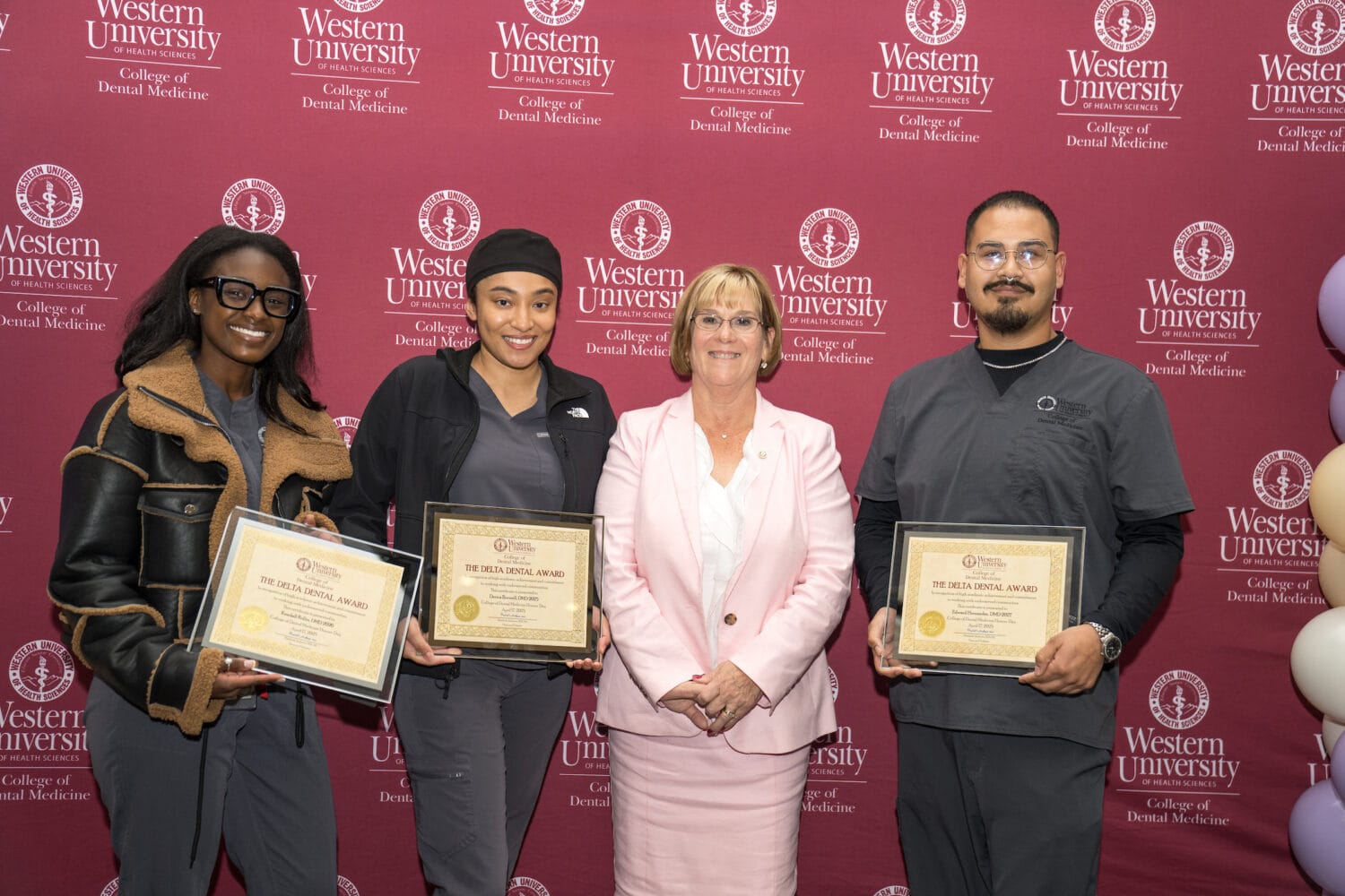 Three individuals holding award certificates stand beside a woman in a white blazer in front of a Western University College of Dental Medicine backdrop.