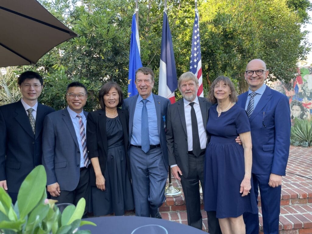 Seven people in formal attire stand together outdoors, smiling for a group photo in front of French and American flags with greenery and a brick patio in the background.