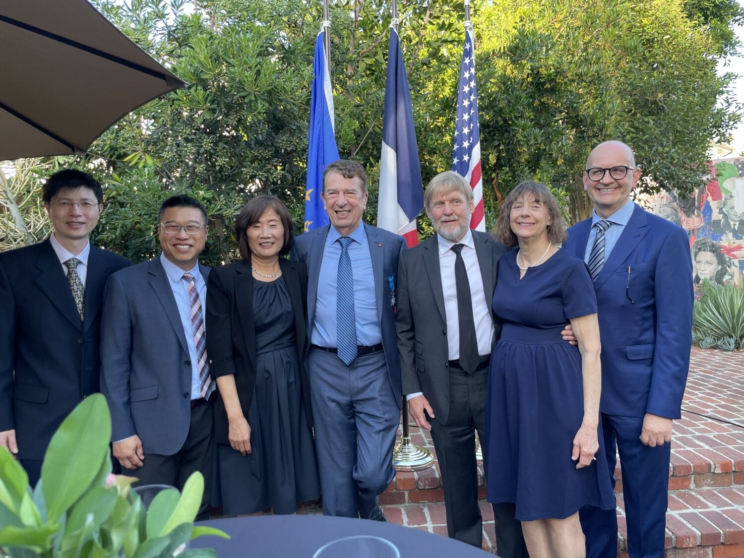 Seven people in formal attire stand together outdoors, smiling for a group photo in front of French and American flags with greenery and a brick patio in the background.