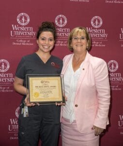 Two women pose in front of a Western University College of Dental Medicine backdrop; one is holding a framed certificate labeled "The Delta Dental Award.