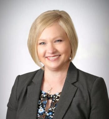 A woman with blonde hair in a bob haircut, wearing a dark blazer and patterned blouse, smiles at the camera against a plain background.