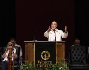A man in a white coat speaks at a podium with the Western University of Health Sciences banner, addressing an audience on stage.