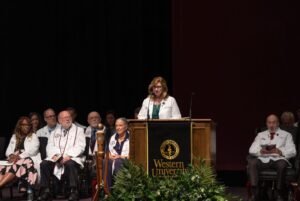 A woman stands at a podium labeled "Western University" addressing an audience, with several people in white coats seated on stage behind her.