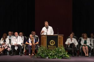 A speaker in a white coat addresses the audience at a podium with a "Western University" banner; several seated individuals in white coats are behind him on stage.