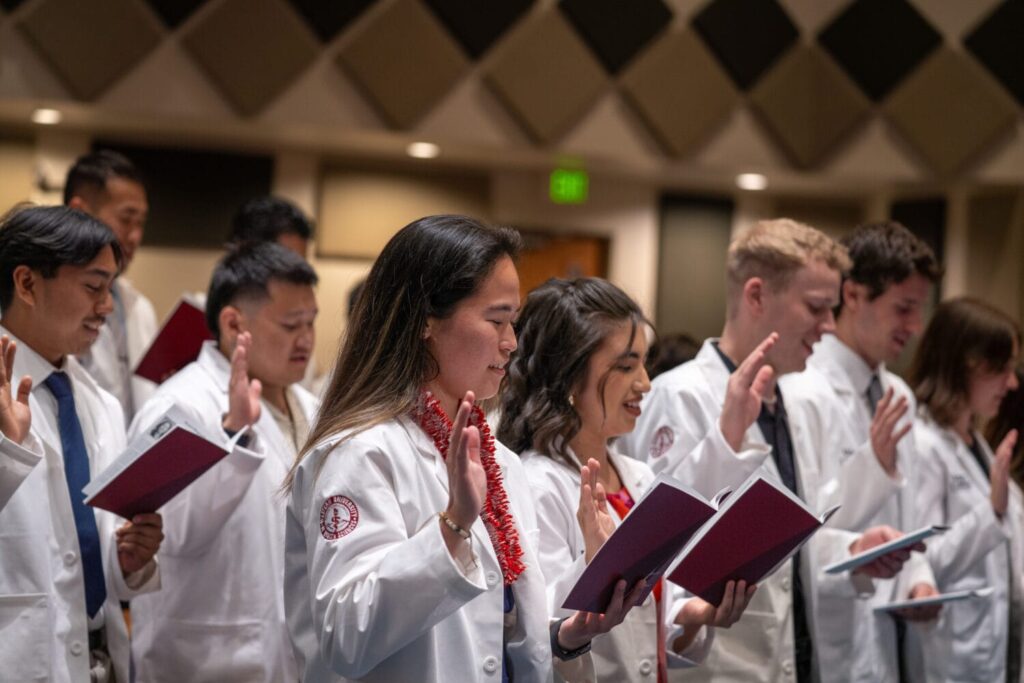 A group of people in white coats hold up their right hands and read from red booklets during a ceremony in an indoor setting.