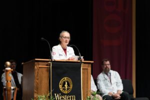 A woman in a white coat speaks at a podium with the Western University logo, while others in white coats sit behind her on stage.
