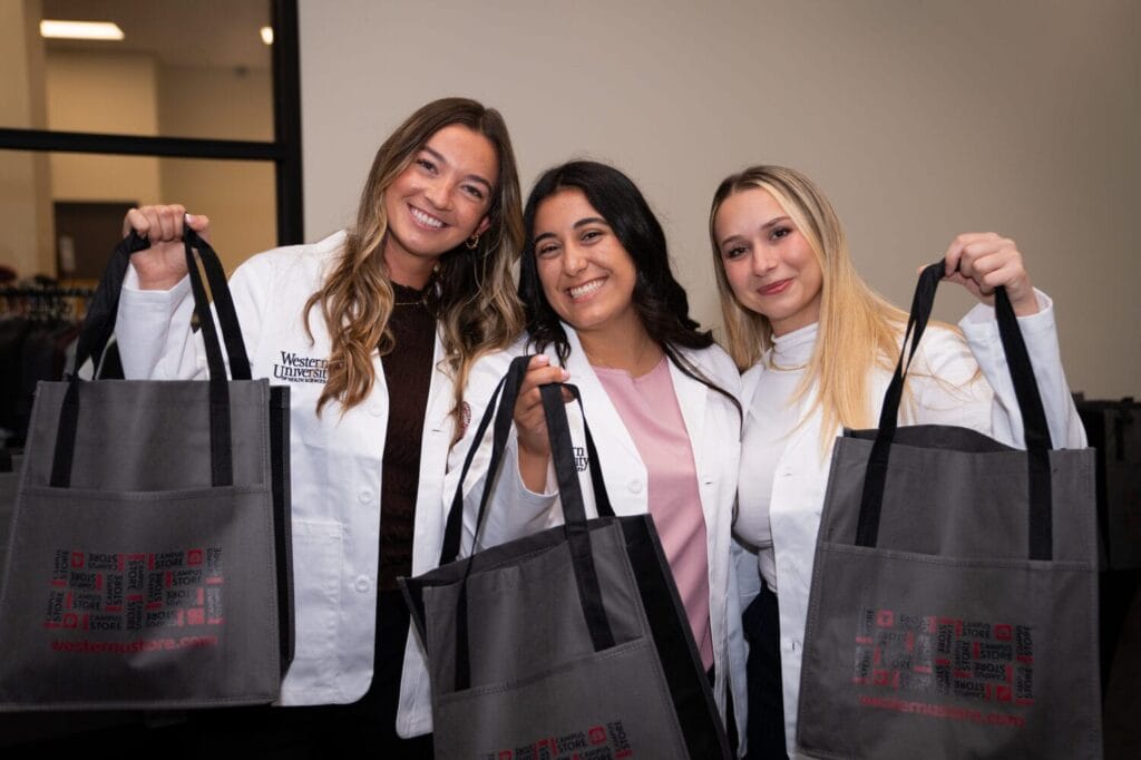 Three women wearing white lab coats hold up black tote bags and smile at the camera in an indoor setting.