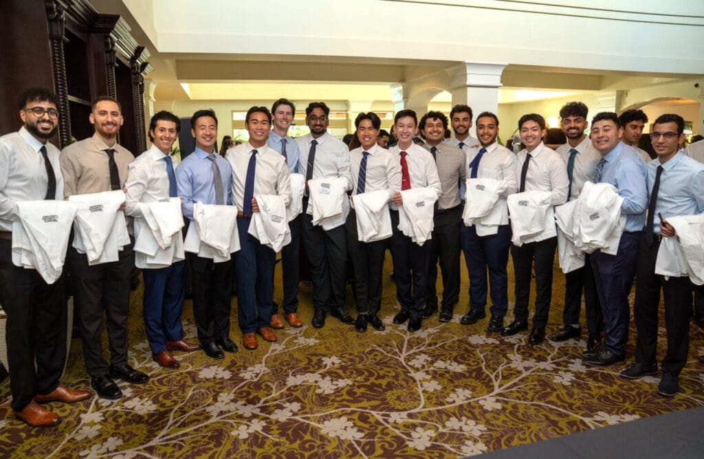 A group of young men in dress shirts and ties stand together indoors, each holding a white coat, posing for a group photo on a patterned carpet.