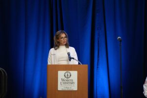 A woman in a white coat stands at a podium labeled "Western University College of Dental Medicine," speaking in front of a blue curtain.