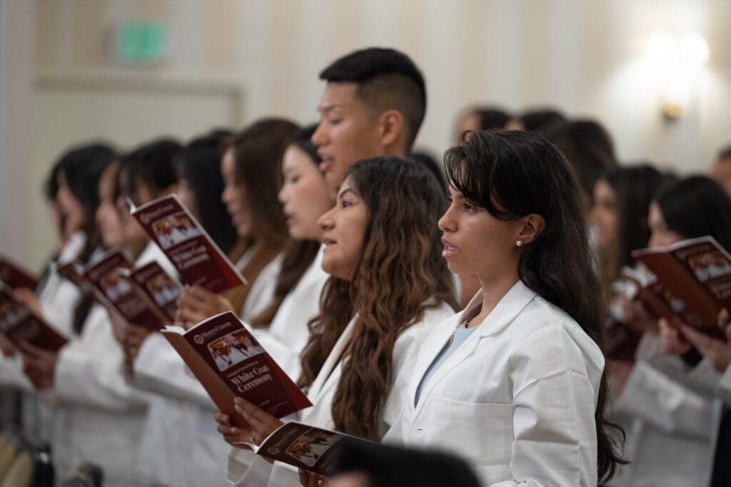 A group of people in white coats stand in rows, holding and reading from booklets during a ceremony.
