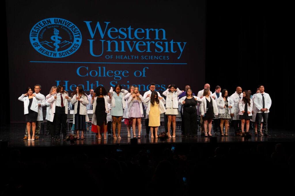A group of people in white coats stand on stage under a projected "Western University College of Health Sciences" sign, participating in a ceremony.