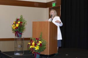 A woman in a white coat speaks at a podium on a stage, flanked by two large floral arrangements.